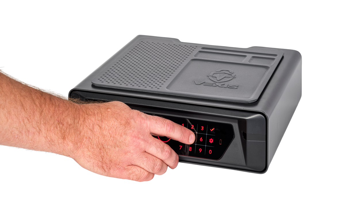 Person's hand entering a PIN code on the red-lit VEXIS touch keypad of the Hornady VEXIS Night Guard bedside safe, demonstrating fast keypad access, compact dark gray safe with rubberized organizer mat on top