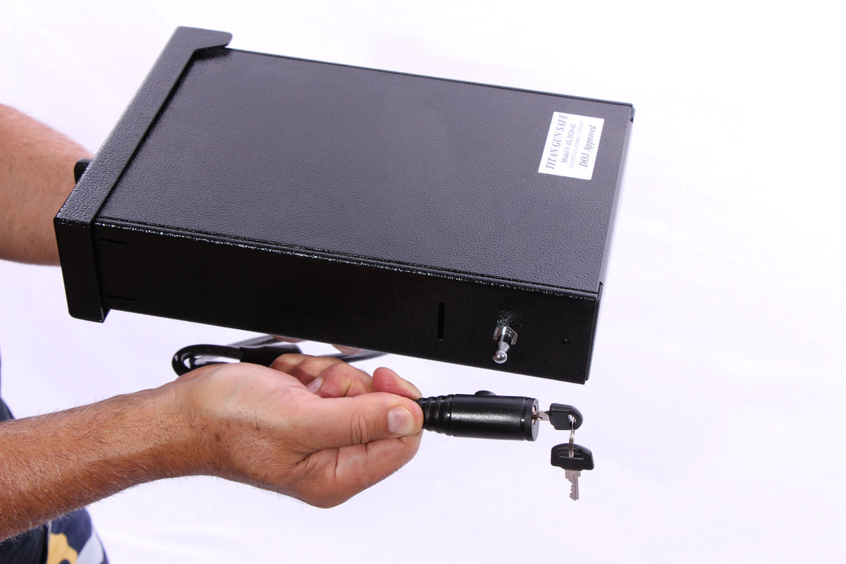 Titan Cable Lock installation demonstration: person holding a Titan Pistol Vault (AS-2020442, California DOJ approved) in one hand and the cable lock's cylindrical mounting pin with keys inserted in the other, showing the pin ready to be attached to the vault's anchor hole