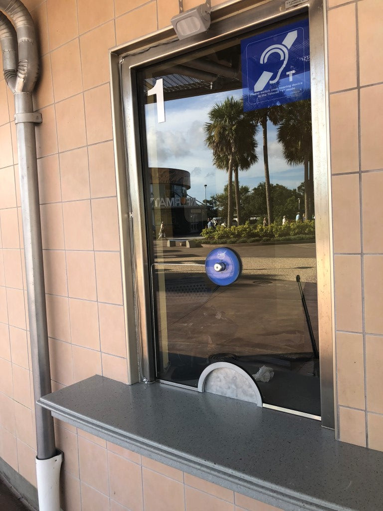 Contacta STS-C100 blue glass-mounted microphone installed at an outdoor ticket window with a hearing loop symbol