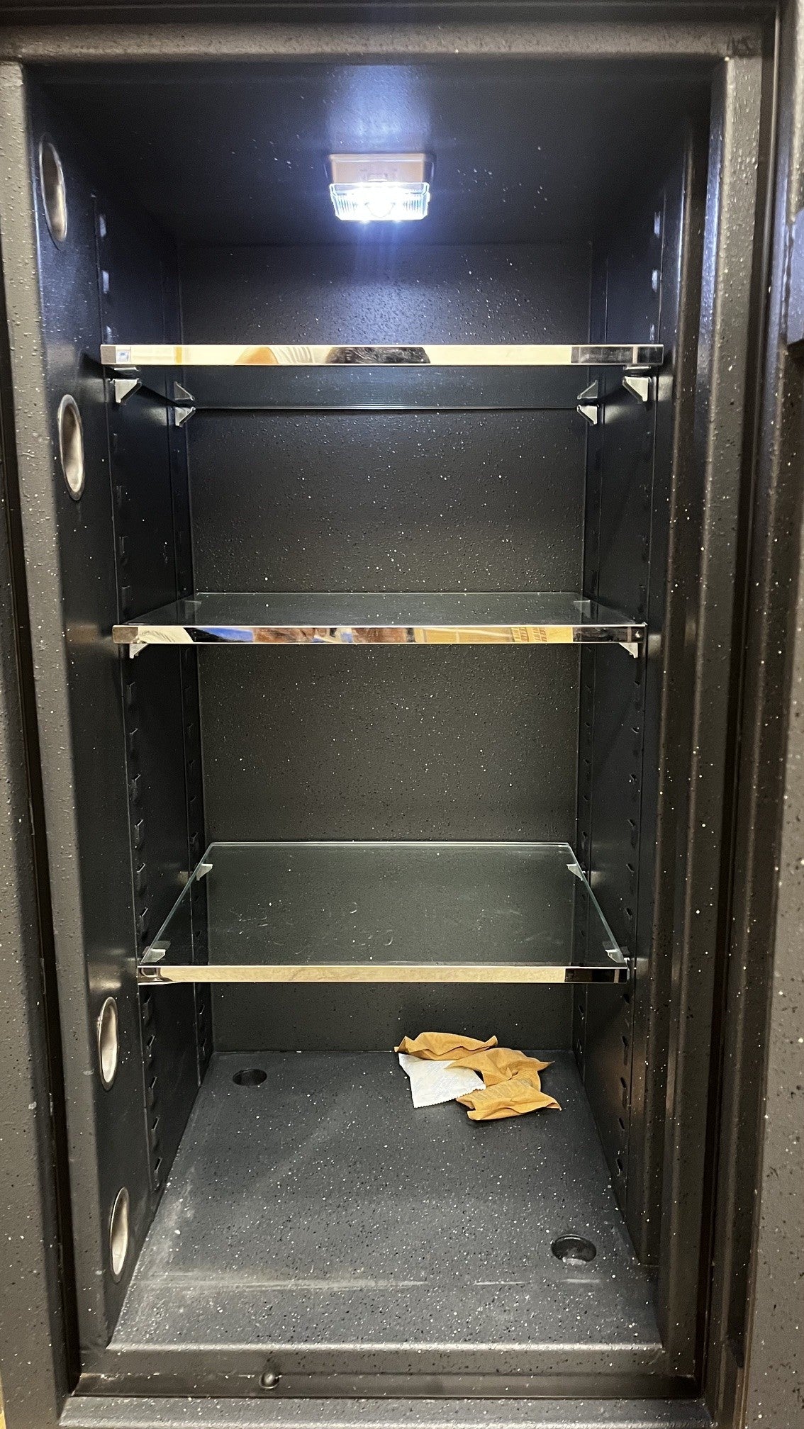 Close-up view of the inside of the safe with the light on. It has three adjustable shelves and two predrilled holes to bolt it down to the floor. It has one hole on the back left corner and one on the right corner. 