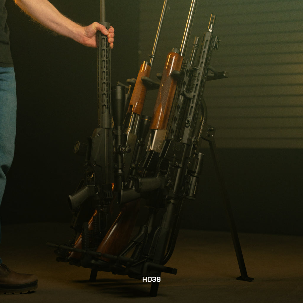 Person holding a rifle against a rack of other rifles in a dimly lit room.