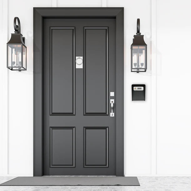 Barska CB14206 wall-mounted key lock box in black installed on the exterior wall to the right of a residential front door, showing discreet placement beside a paneled entry door and lantern-style porch lights
