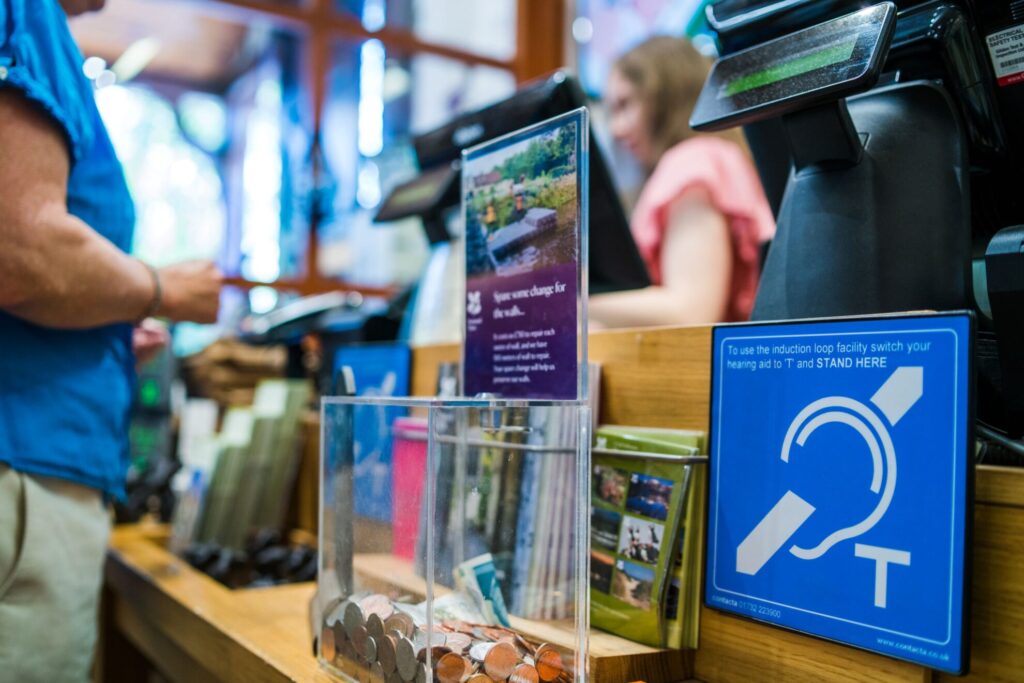 Contacta IL-AE97 T-coil sign on a museum shop counter next to a donation box and merchandise, staff at the register behind