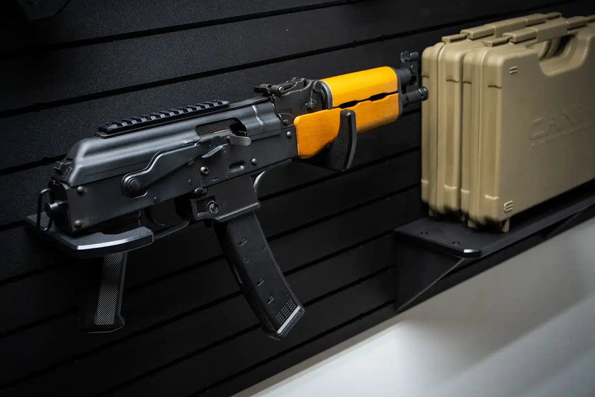 AK-platform pistol with orange wooden furniture, folding stock, and 30-round magazine displayed horizontally on a Tactical Walls MWHRACKRS ModWall rack pair next to a tan hard case on a matching ModWall shelf accessory