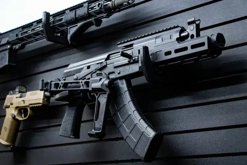 Two modern rifles displayed on a Tactical Walls ModWall panel: an AR-platform rifle at top and a folded-stock AK variant with black magazine below, plus a tan Sig Sauer pistol mounted underneath, all using Tactical Walls ModWall panel accessories