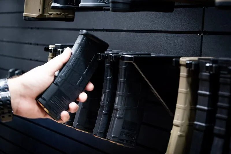 Hand holding a black rifle magazine in front of a wall of similar magazines.