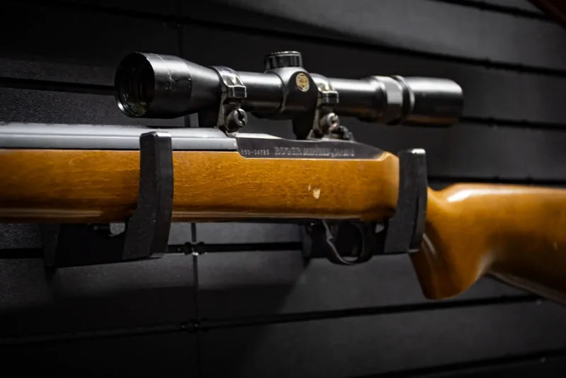 Bolt-action rifle with wooden stock and mounted scope resting horizontally on two Tactical Walls MWHRACKRS ModWall rifle racks installed on a black ModWall panel, showing the parallel-mount configuration holding a long gun