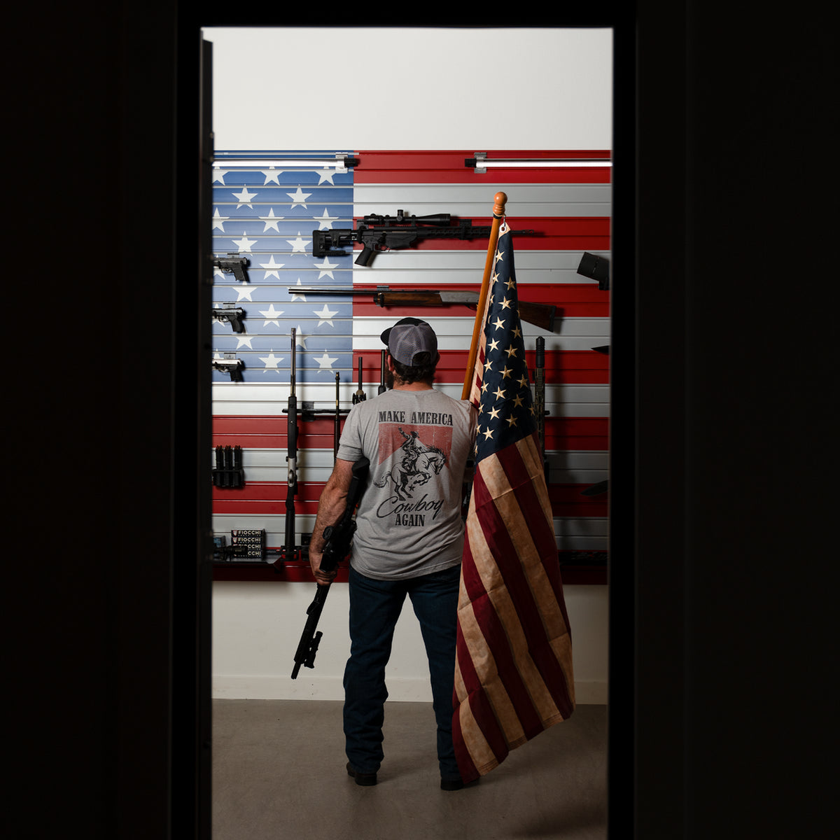 Person holding an American flag and a rifle in front of a wall with American flags and rifles.