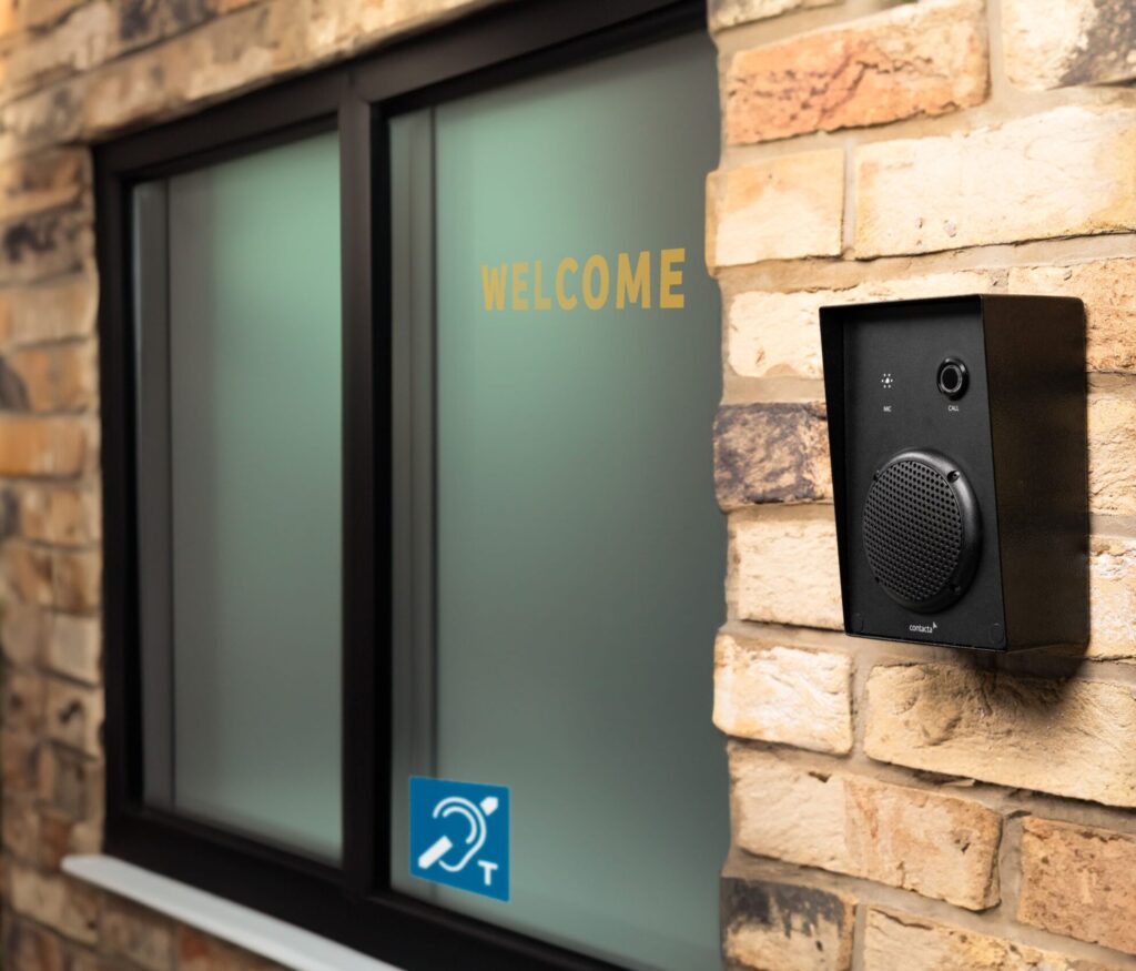 K009-IP outdoor call unit installed on a brick exterior wall beside a welcome window with a T-coil hearing loop symbol