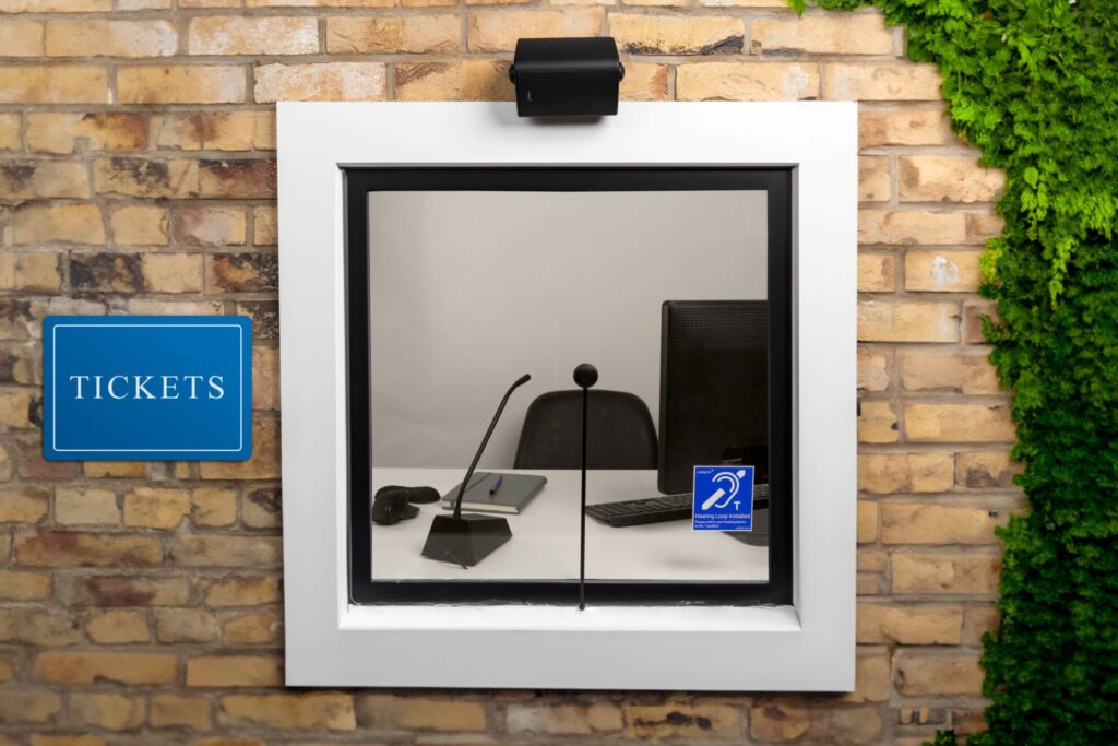 STS-K080-IP overhead speaker mounted above a brick ticket window with a hearing loop symbol and stem mic inside