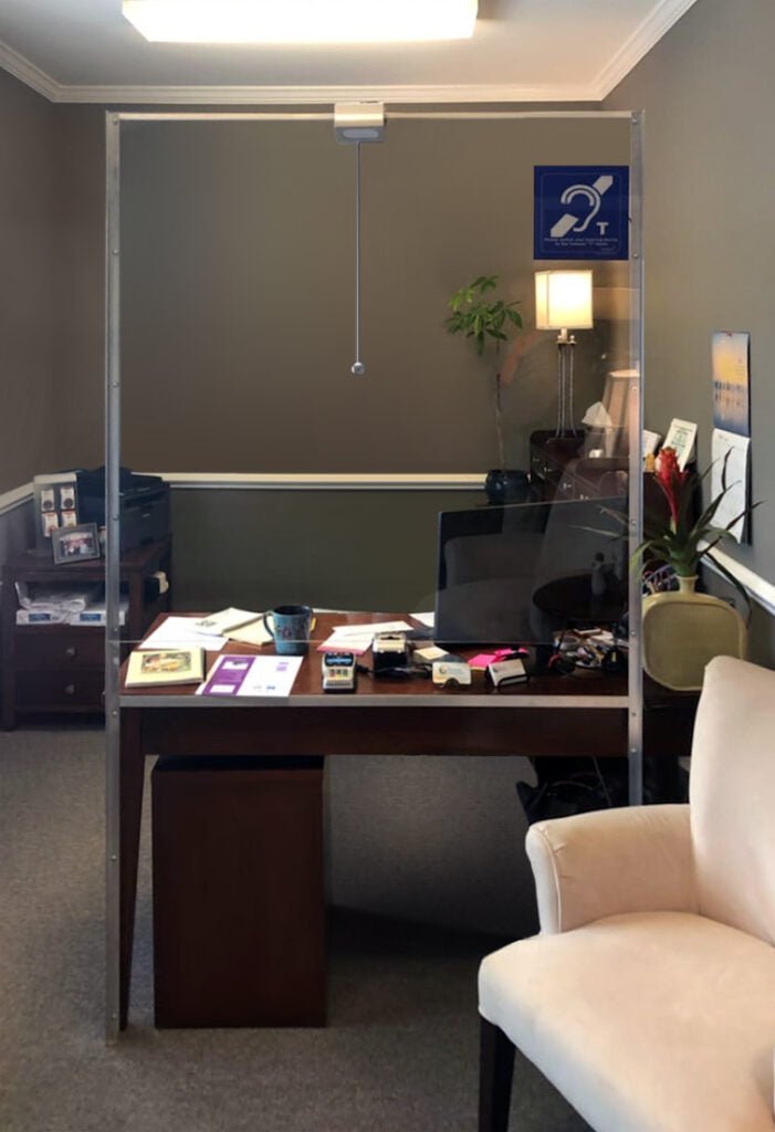 Contacta STS-K064 installed at a reception counter with the STS-S23 overhead speaker suspended above the glass barrier, out of arm's reach, and a blue hearing loop T-coil sign posted on the barrier for visitors using hearing aids
