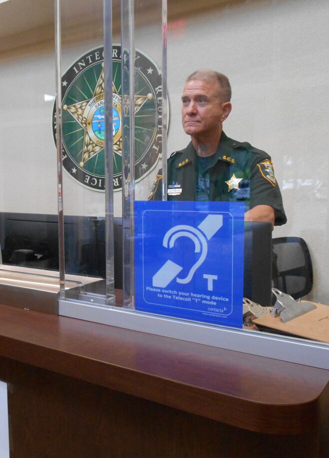 Contacta IL-AE98 mounted on the glass at a sheriff's office reception counter, with a deputy on the staff side of the barrier