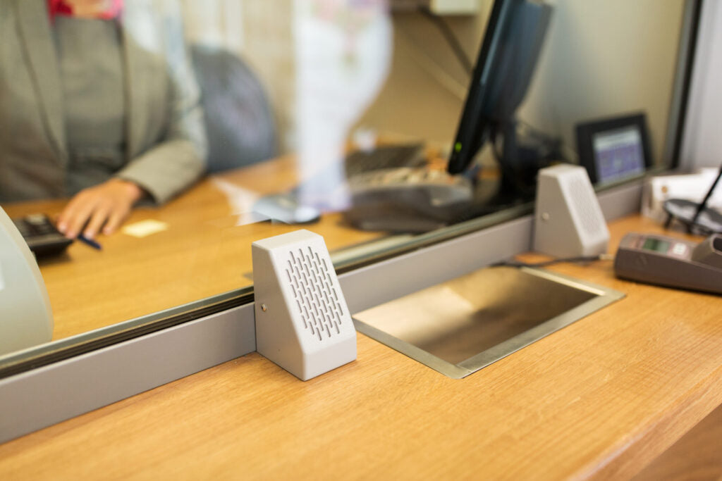 Close-up of the STS-K002 grey customer-side surface-mounted unit installed on a wooden counter beside a glass barrier