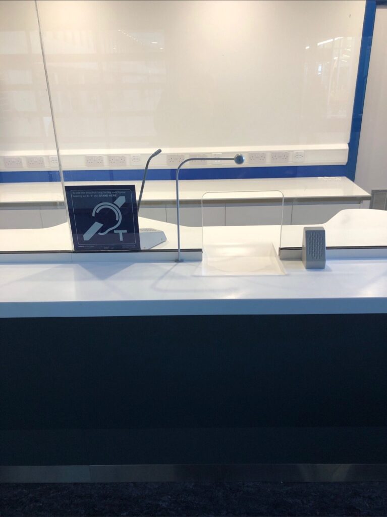 Close-up of the STS-K058 curved-tubing microphone at a service counter with security glass and hearing loop symbol