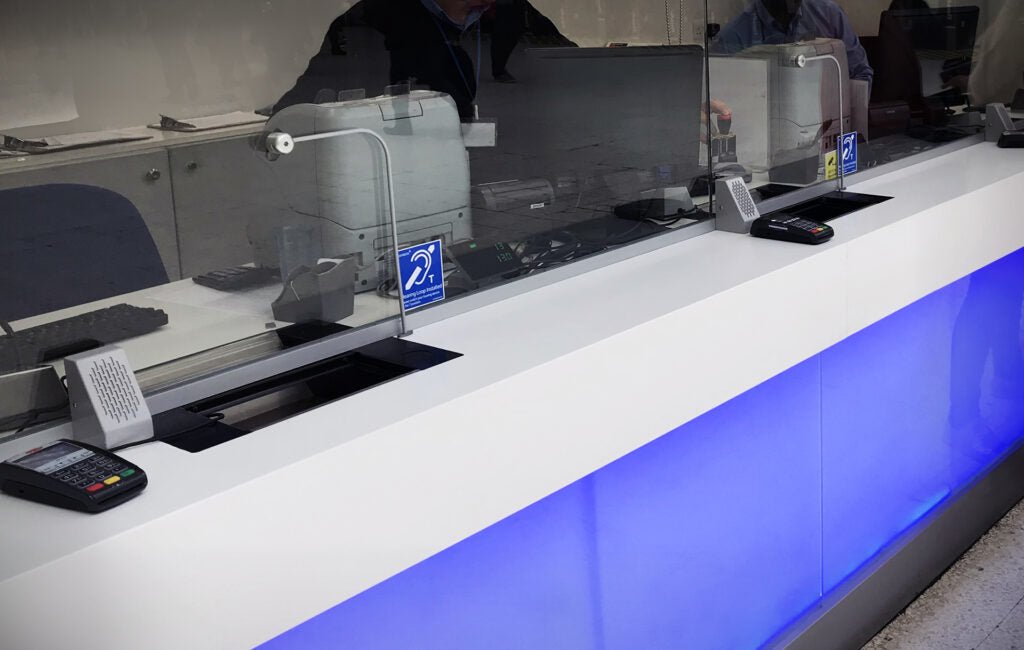Contacta STS-K058 installed at a bank counter with security glass, curved-tubing microphone, and hearing loop symbol