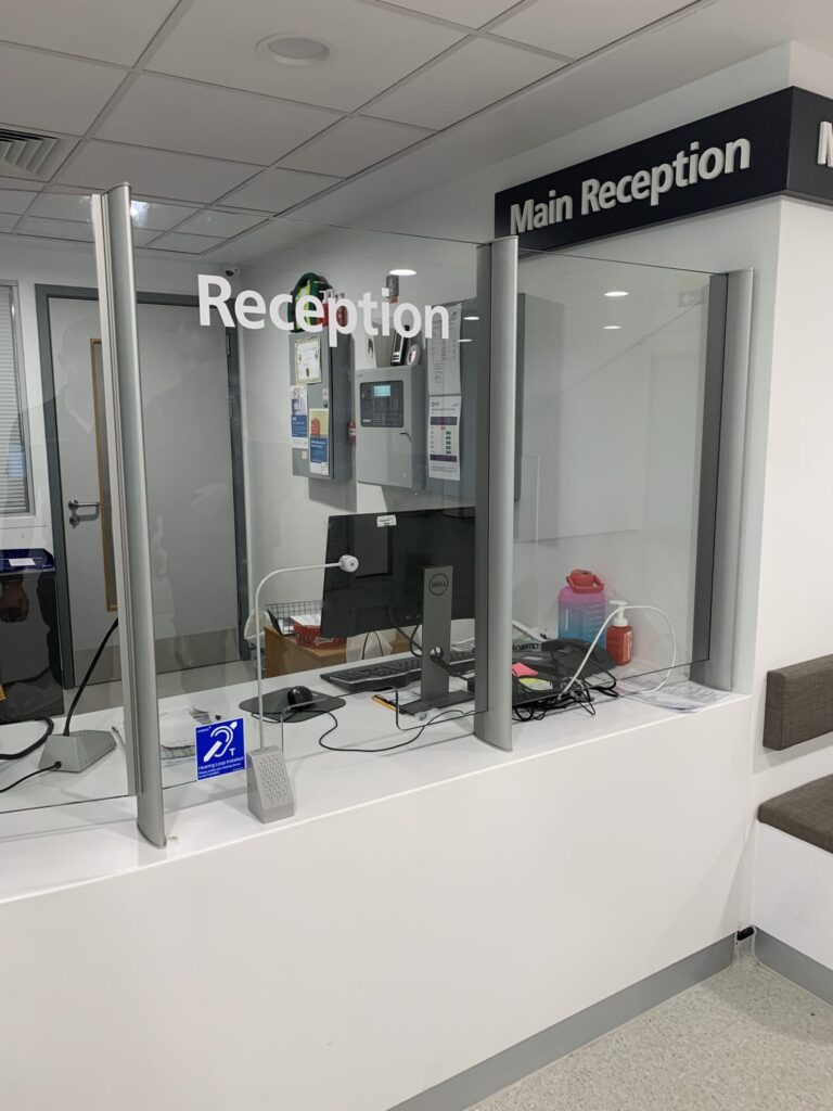 Contacta STS-K058 curved-tubing microphone installed at a main reception counter with security glass and hearing loop signage