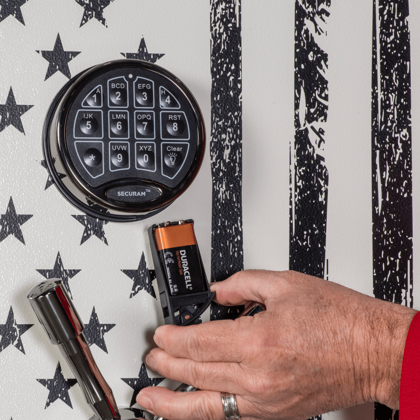 Close-up of the Winchester Ranger 26 Americana Securam electronic keypad set against the American flag door graphic, with a hand replacing the 9-volt battery next to the chrome vault handle